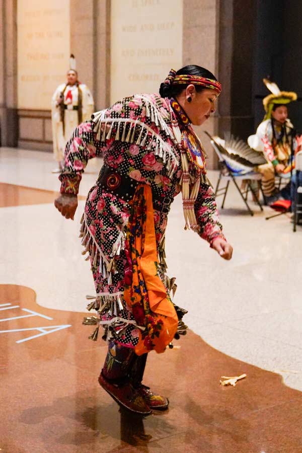 a Native American woman wearing a pink and orange jingle dress during a dance held in the Bullock Museum Grand Lobby
