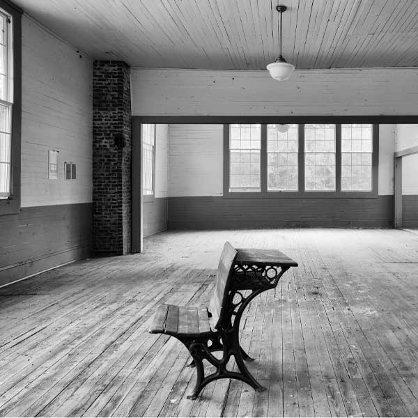 a black and white photo of an empty classroom with a single old wooden desk facing the window