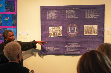 a Black woman instructing a class and pointing to a quilt on a wall