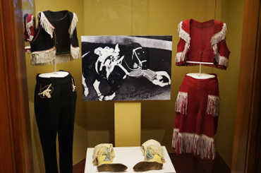 a museum display case with two cowgirl outfits and a photo of a cowgirl riding a horse.