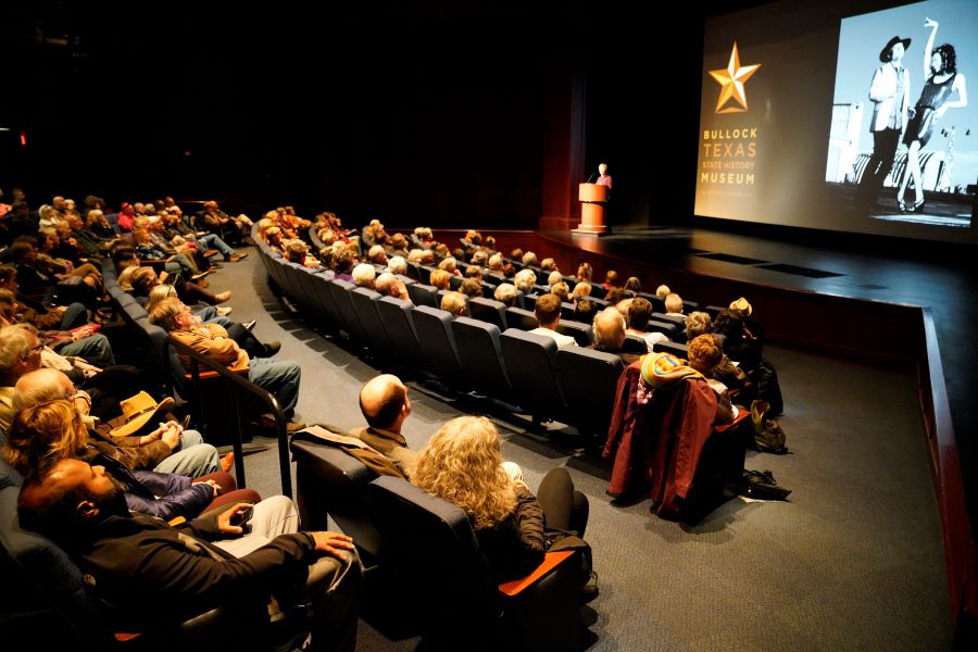 people seated in a theater listening to a speaker on stage 